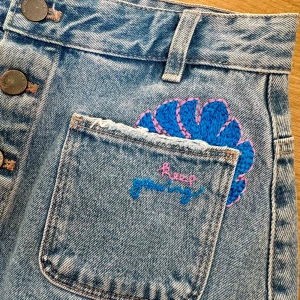 Jeanskjol med broderade detaljer - Säljer en superfin jeanskjol med coola broderade detaljer. Kjolen har en knappknäppning framtill och två fickor på baksidan. På framsidan finns det broderade löv i blått och rosa samt texten 'keep growing'. Perfekt för en avslappnad och trendig look!