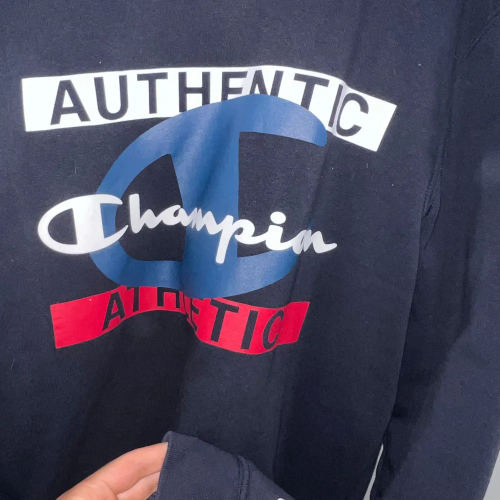 Säljer en snygg mörkblå tröja från Champion i storlek S. Den har ett stort tryck på framsidan med texten 'Authentic Athletic' i vitt och rött. Tröjan är långärmad och perfekt för en casual look. Passar bra till både jeans och träningsbyxor!. Tröjor & Koftor.