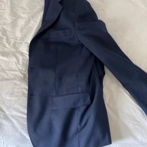 Mörkblå kostym från D+LJ - Säljer en stilren mörkblå kostym från D+LJ. Kavajen har en klassisk passform med två knappar och fickor fram. Byxorna matchar perfekt och har en elegant skärning. Perfekt för både formella tillfällen och fest.