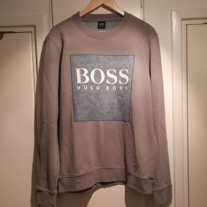 Säljer en stilren grå sweatshirt från Hugo Boss. Den har en stor, blå fyrkant med BOSS-loggan i vitt på framsidan. Tröjan är långärmad och har en normal passform, perfekt för både vardag och avslappnade tillfällen. Materialet känns mjukt och bekvämt.