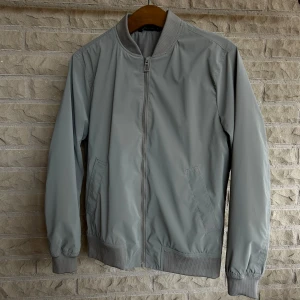 Grå bomberjacka - Säljer en stilren grå bomberjacka i nyskick. Jackan har en dragkedja framtill och ribbade muddar vid ärmslut och nederkant. Perfekt för vår och höst!