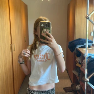 Vit t-shirt från Juicy Couture - Säljer en superfin vit t-shirt från Juicy Couture med ett coolt orange tryck på framsidan. Den är kortärmad och har en croppad passform, perfekt för en avslappnad stil. Passar bra till både jeans och kjol! märket är inte förstörd, de är meningen! säljs för jag inte använder den tyvärr den är i bra skick och skön. köptes för ungefär 800kr💞