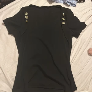 Svart topp med guldknappar från SHEIN - Säljer en snygg svart topp från SHEIN i storlek S. Den har korta ärmar och är dekorerad med stiliga guldknappar på axlarna. Perfekt för en kväll ute eller en festlig tillställning. Toppen är i bra skick och redo för nya äventyr!