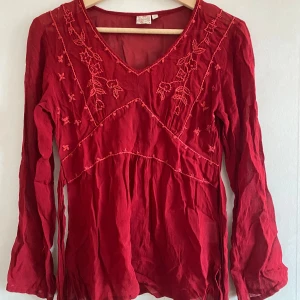 Röd blus från Anokhi - Säljer en superfin röd blus från Indiska. Den har v-ringning och långa ärmar med vackra broderade detaljer. Blusen är i lätt material, perfekt för både vardag och fest. Den har också ett knytband i midjan för en snygg passform. Den är inte helt genomskinlig och har inga defekter💓