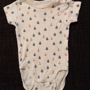 Body strl. 50/56 - Vit body med marint motiv (båtar och ankare) i storlek 50/56 från Åhlens. I fint skick, inga hål eller fläckar men något blekare än ny.  Tvättad i parfymfritt tvättmedel och utan sköljmedel.