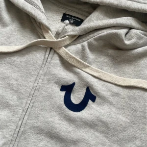 True Religion zip up - grå truey hoodie me backprint, skön zipup. sparsamt använd, inga skavanker över huvud taget. storlek M-L beroende på hur du vill att den ska sitta.