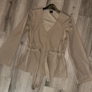 Gina tröja  - I fint material som glänser lite (svårt att få till på bild, det är inte som satin men glänser lite). I fint skick, man spänner åt i midjan och knyter när man har den på. Grå/beige färg som också är svår att fånga på bild 