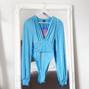 Ljusblå body XS urringad  - Färg: ljusblå.  Strl: XS.   Snygg body med skimrigt tyg. Elastiskt och skönt material. Djup urringning framtill. Knäpps med dolda knappar mellan benen. -OBS. Helt ny!  Kommer ej till användning & stor garderobsrensning pågår!