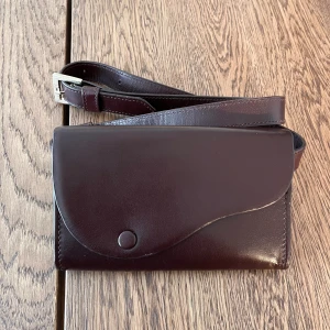 Midjeväska - Midjeväska från Wera i mörkbrunt/burgundy. Den har några små skav från normal användning, men inga större defekter. Det finns ett fack med dragkedja och det finns även fack för att förvara bankkort och liknande. 