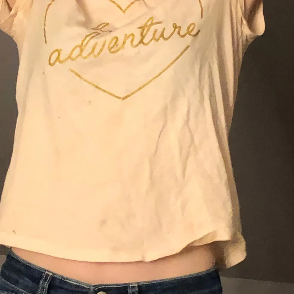 En ljusrosa kortärmad med ett hjärta i guld och inne i hjärtat står de hello adventure . T-shirts.