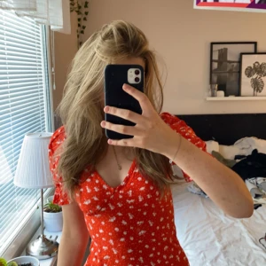 Röd blommig klänning - En röd klänning med vita blommor. Klänningen har korta ärmar och en v-ringad hals. Den är figursydd och har en omlott-design. Har en dragkedja på sidan som gör det lättare att ta på sig den