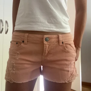 Lågmidjade jeansshorts - JÄTTESNYGGA aprikos-rosa lågmidjade jeansshorts med fina spetsdetaljer på sidorna! Kan ej hitta några defekter på dem, som i nyskick. Priset kan diskuteras💕