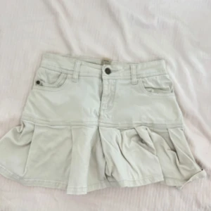 Beige kjol med volanger - En beige kjol med volanger och fickor fram och bak. Kjolen har en knapp och dragkedja framtill samt bälteshällor.