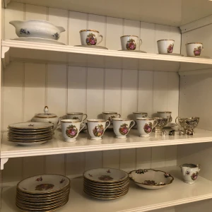 Porslinssamling med blommotiv - En samling av porslin med blommotiv. Samlingen inkluderar koppar, fat, skålar och en såssnipa. Porslinet är dekorerat med blommor i rosa, lila och gröna nyanser samt har guldkant.