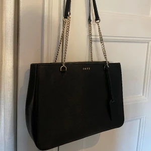 DKNY väska svart - Klassisk väska från DKNY med gulddetaljer. Två innerfack varav ett med dragkedja. Knappt några tecken på användning och nypris ca 2500 kr.
