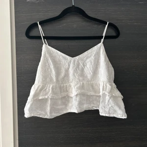 Linne - Vit spets topp med volanger. Toppen är i en fin spets med små blommor och har en avslappnad passform. Den är perfekt för varma dagar eller för att klä upp en enkel outfit.