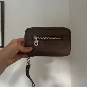 Brun plånbok med dragkedja - En kompakt brun plånbok med dragkedja och avtagbar handledsrem. Plånboken har flera fack för kort och en liten ficka med dragkedja inuti.