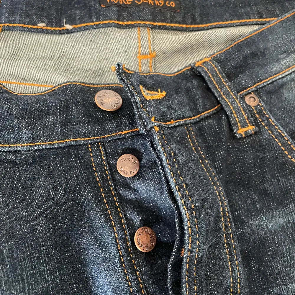 Ett par mörkblå jeans från Nudie Jeans med modellen Lean Dean Troubled Sea. Jeansen har en knappgylf och orange sömmar. Passar storlekarna M/L.. Jeans & Byxor.