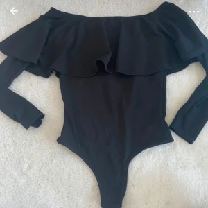 Svart off-shoulder body - En elegant svart off-shoulder body med långa ärmar och volangdetaljer runt halsen. Perfekt för festliga tillfällen.