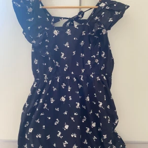 Svart klänning med vita blommor på - En lång svart klänning me vita blommor på använd 1 gång. Har en jätte fin roset bak. Från Gina 