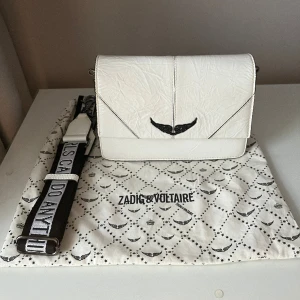 Vit väska Zadig & Voltaire - Säljer en helt slutsåld väska från Zadig & Voltaire, självklart äkta. Väskan är i bra skick och även bandet. Just bandet användes aldrig eftersom jag bytte ut det mot ett eget. Dustbag tillkommer. Väskans mått är: bredd 21, höjd 15 och djup 12cm. 