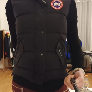 Äkta Canada Goose  - Damväst från Canada Goose med två snedställda framfickor, en innerficka och broderad applikation på vänster bröst. Västen stängs med dragkedja och tryckknappar.  Stl S/P. Sitter bra på mig som har stl 36/38.  Fint skick!  Åkta