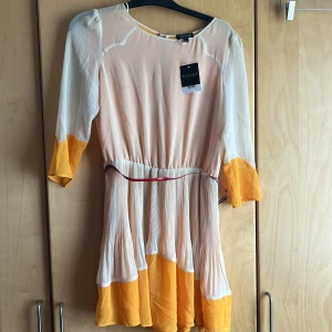 Tvåfärgad fin klänning  - Splitterny med prislappen på, röd plastig skärp medföljer det går bra att ta bort skärp. Tvåfärgad skir tyg ej genomskinlig, veckad i kjoldelen . Creme/orange/gul i färg 