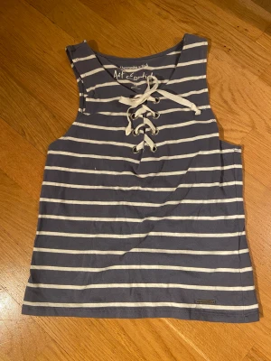 Randigt linne - Ett blått/grått och vitt randigt gulligt linne med vita snören. Inte min stil längre men såå söt❤️Passar många storlekar, den är liten men stretchig!