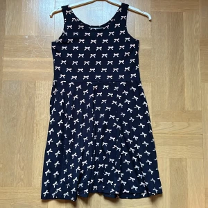 Mörkblå klänning med rosett-mönster - En mörkblå klänning med ett mönster av ljusrosa rosetter. Klänningen är ärmlös och har en normal passform. Den är knälång och tillverkad i ett mjukt och bekvämt material.