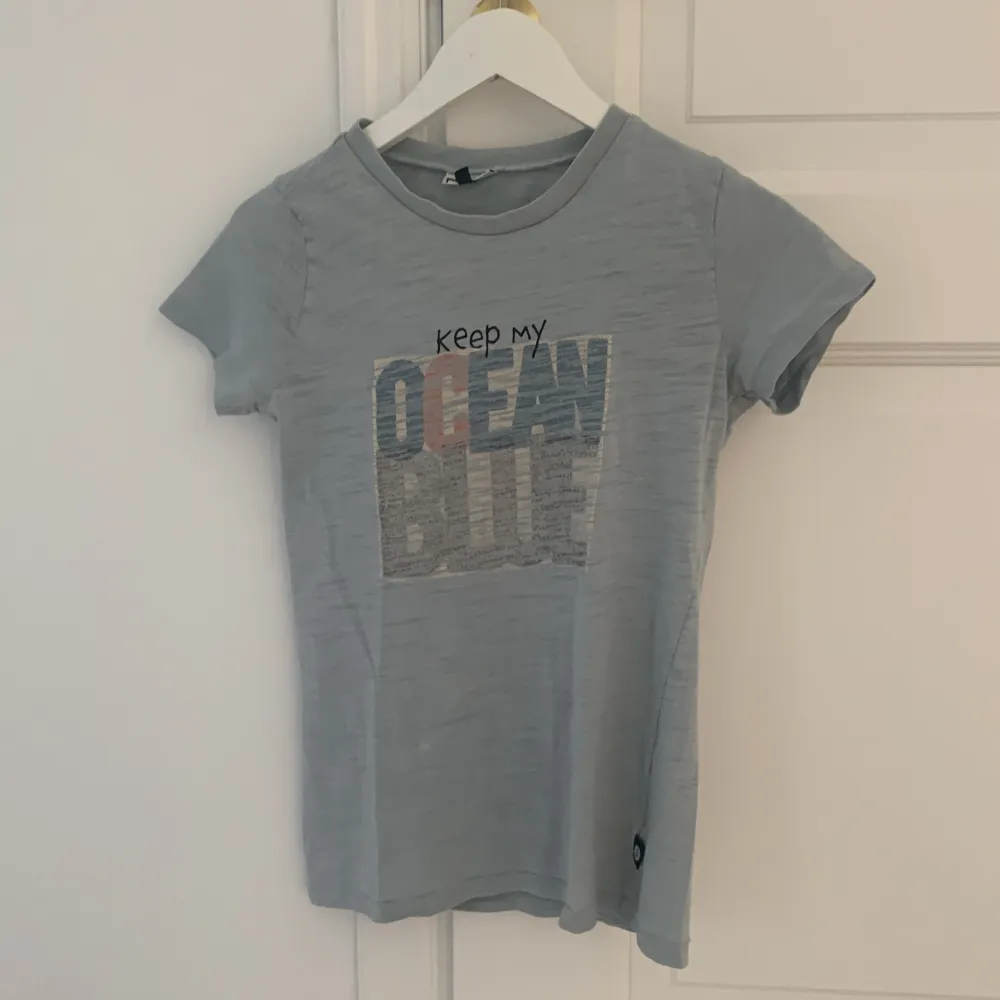 En grå t-shirt med texten 'Keep my ocean blue' tryckt på framsidan. T-shirten har korta ärmar och en normal passform.. T-shirts.