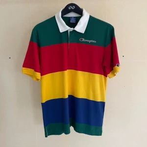 Flerfärgad pikétröja från Champion - aEn färgglad pikétröja från Champion med breda horisontella ränder i grönt, rött, gult och blått. Tröjan har en vit krage och korta ärmar. Champion-logotypen är broderad på bröstet och på vänster ärm.