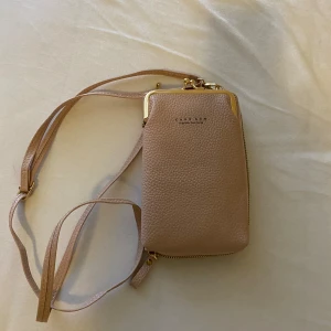 Rosa oanvänd CARR KEN väska  - Den här lilla crossbody-handväskan är tillverkad av PU-läder. Väskan är uppdelad i två stora fickor, den ena stängs med dragkedja och den andra stängs med lock.   En praktisk och fin väska som fyller alla funktioner i en plånbok med olika fickor. 