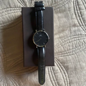 Daniel Wellington klocka - Klocka från Daniel Wellington i fint skick, enbart enstaka ytliga repor på uret. Svart läderband och ur, samt silvriga detaljer.