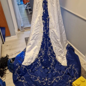 Brudklänning - ej använd - Brudklänning i vitt och marinblått o silver satin med släp. Broderier på. Tyvärr är den liten skrynklig men detta går att ånga bort. Den har varit i paket. Köpt från internet. Storlek 18W Dam plus storlek Large Storleksguide i bilderna.  Built in bra