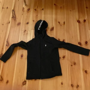 Svart jacka från Peak Performance - Säljer en svart jacka från Peak Performance. Den har en huva med texten 'PERFORMANCE' och dragkedja framtill. Jackan är i bra skick och perfekt för både vår och höst. Den är gjord av ett mjukt och bekvämt material.