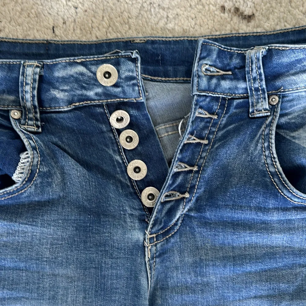 Säljer ett par snygga blå jeansshorts från Place du Jour i storlek 36. De är högmidjade och har en cool knappdetalj framtill. Perfekta för sommaren! Shortsen är i nyskick och har en normal passform. Materialet är mestadels bomull med lite elastan för extra komfort.. Shorts.