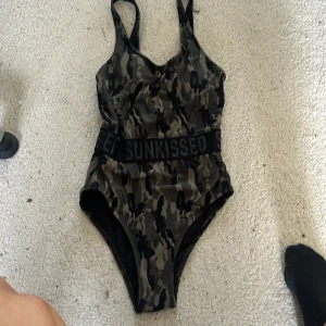 Kamouflagemönstrad baddräkt från Sunkissed - Säljer en supercool kamouflagemönstrad baddräkt från Sunkissed. Den har texten 'Sunkissed' tryckt flera gånger på banden och en snygg öppen rygg. Perfekt för sommarens bad och strandhäng! Baddräkten är i nyskick och har en tight passform.