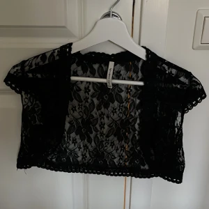 Svart spetsbolero - Säljer en svart spetsbolero med blommönster. Den är kortärmad och har en fin, detaljerad spetskant. Perfekt för att lägga till en elegant touch till din outfit. Passar bra till fest eller andra speciella tillfällen. superbra skick
