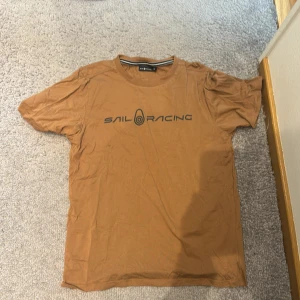 Brun t-shirt från Sail Racing - Säljer en brun t-shirt från Sail Racing. Den är i bomull och har en normal passform. T-shirten har korta ärmar och ett tryck med märkesnamnet på framsidan. Perfekt för vardagsbruk eller träning.