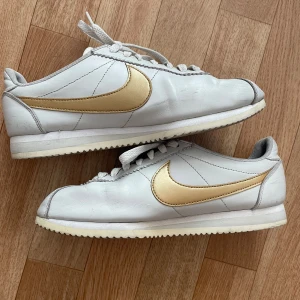 Vita Nike Cortez sneakers med gulddetaljer - Ett par vita Nike Cortez sneakers med gulddetaljer🤗Skorna har en vit bas med en guld Nike-logga på sidan. De är storlek 40,5 men jag har storlek 39- 40 o de funkar bra de me.