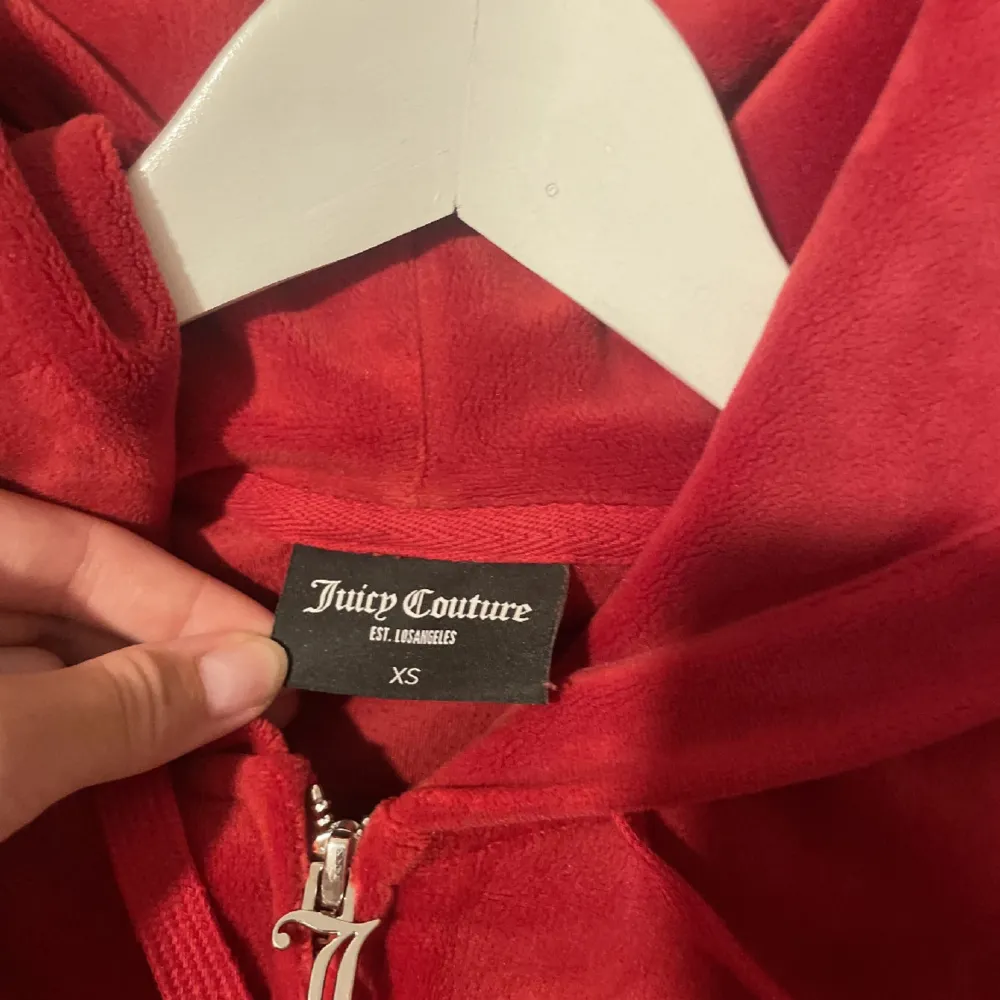 Säljer en snygg röd hoodie från Juicy Couture i storlek XS. Den är gjord av mjukt material och har en dragkedja framtill. Perfekt för en avslappnad stil eller för att hålla sig varm under kyliga dagar. Hoodien har långa ärmar och en praktisk huva med snören.. Hoodies.