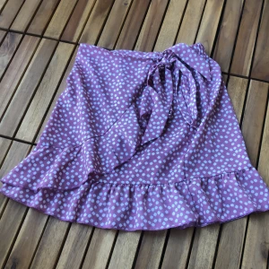 Lila kjol från SHEIN - En rosa-lila kjol från SHEIN med vita blommor. Kjolen har en volangdetalj längst ner, resår i midjan på baksidan och ett knytband i midjan. Tillverkad av 100% polyester.