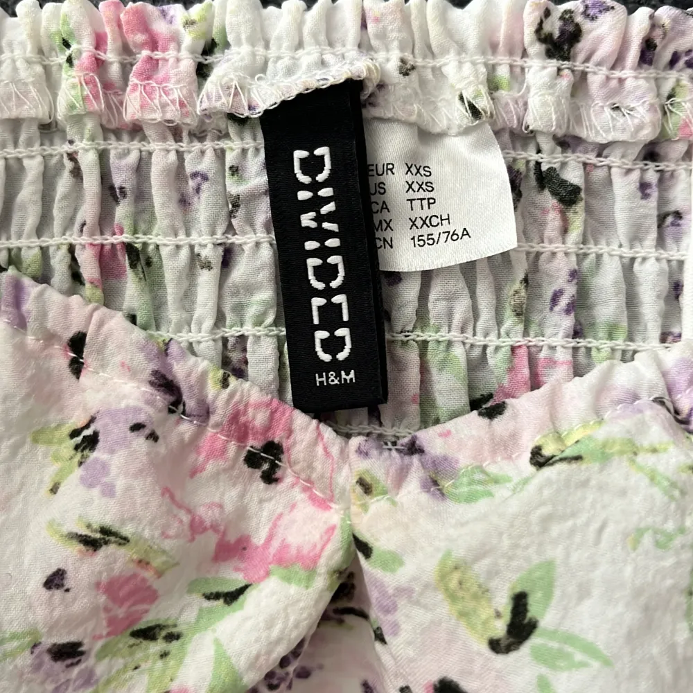 En söt blommig klänning från H&M med puffärmar och en smockad överdel. Klänningen har en vit bas med ett mönster av rosa, lila och gröna blommor. Den är kort och har en tight passform.. Mekot.