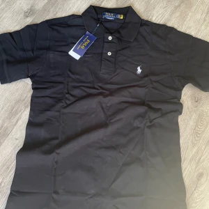 Svart pikétröja från Polo Ralph Lauren - En svart pikétröja från Polo Ralph Lauren i storlek M. Tröjan har korta ärmar och en klassisk krage med knappar. Den är dekorerad med en broderad logotyp på bröstet. tags ej kvar