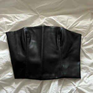 Svart korsett från Gina Tricot - En stilren svart korsett från Gina Tricot i storlek XS. Korsetten har en dragkedja på baksidan och är tillverkad i ett syntetmaterial.