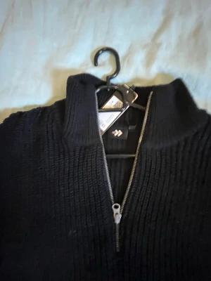 Svart stickad halvzip-tröja - Säljer en svart stickad tröja från lager 157  med halvzip och hög krage. Tröjan har lång ärm och är ribbstickad vilket ger en snygg struktur. Perfekt för lager på lager och enkel att matcha med olika outfits.
