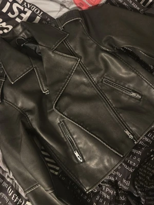 Svart biker skinnjacka med dragkedjor - Snygg svart skinnjacka i klassisk bikerstil med bred krage och flera dragkedjor framtill. Jackan har en cool, stilren look och passar perfekt till streetwear eller en edgy outfit. Inga synliga loggor eller märken.