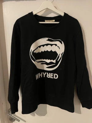 Svart sweatshirt från Whyred - Sällsynt svart sweatshirt från Whyred med ett stort vitt tryck. Tröjan har rund halsringning, långa ärmar och ribbade muddar. Perfekt för dig som gillar streetwear och grafiska prints.