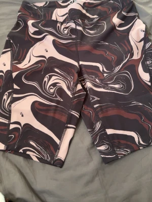 Mönstrade träningsshorts i svart och beige - Säljer ett par snygga träningsshorts med abstrakt mönster i svart, beige och brunt. Shortsen har en kroppsnära passform och är tillverkade i ett stretchigt funktionsmaterial som passar perfekt för gym eller löpning.