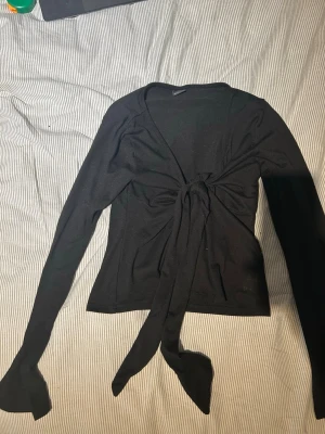 Svart omlottopp med knytband - Snygg svart omlottopp med långa ärmar och knytband framtill som ger en cool och trendig look. Toppen har en figurnära passform och är tillverkad i mjukt material som känns skönt mot huden. Perfekt att styla med jeans eller kjol.